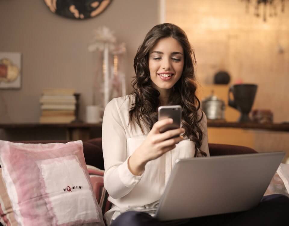 woman-sitting-on-sofa-while-looking-at-phone-with-laptop-on-920382